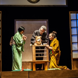 Wnętrze domu. Z obu stron znajdują się parawany przypominające duże okna. W centrum przy stoliku znajdują się trzy osoby. Patrzą z uwagą i zachwytem na siedzącego na stoliku szczeniaka o blond sierści, białym pyszczku i brzuchu. Po lewej stoi kobieta w jasnozielonym kimonie, po prawej klęczy dziewczyna w żółtym kimonie, a na środku stoi mężczyzna w czarnym kimonie i pochyla się nad pieskiem.