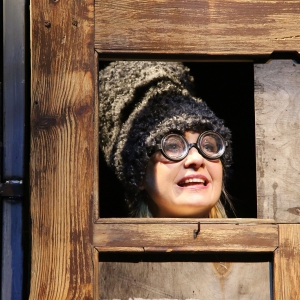 Zdjęcie przedstawia twarz aktorki wyglądającej przez kwadratowe okienko w drewnianej ścianie lub drzwiach.  Ma otwarte w uśmiechu usta, wielką futrzaną czapkę i grube szkła w okularach.