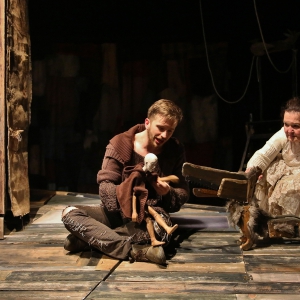 Na pierwszym planie na zniszczonej podłodze z znajduje się dwójka aktorów, po lewej stronie widzimy stare drewniane drzwi bez klamki. Aktor siedzi po turecku i na kolanie trzyma niedużą drewnianą lalkę teatralną, przedstawiającą człowieka z ogoloną g